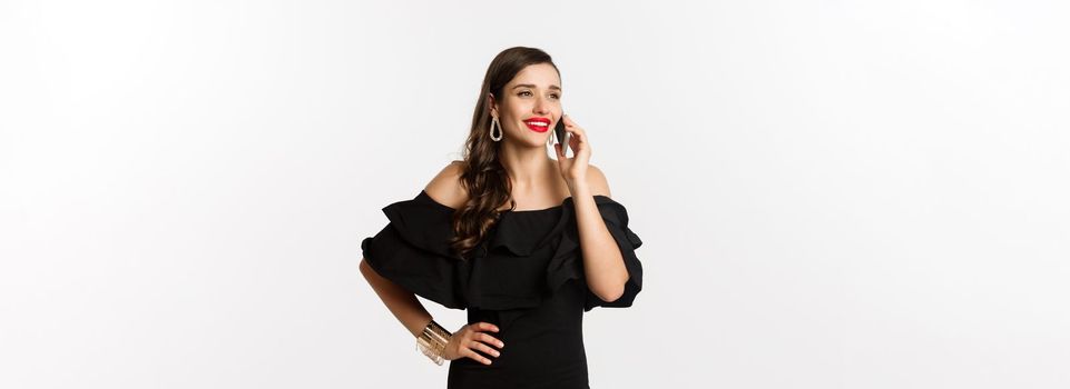 Successful young woman in black dress, red lipstick and makeup, talking on mobile phone and smiling, standing over white background.