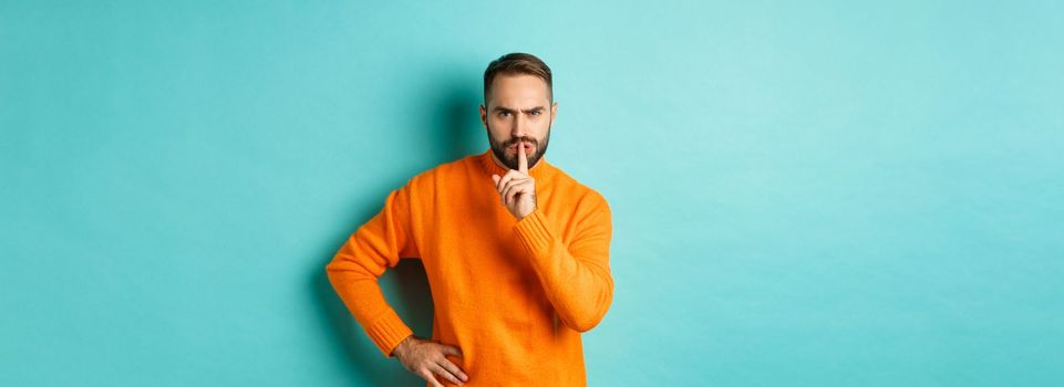 Angry bearded guy telling be quiet, hushing and frowning displeased, shut up gesture, standing over turquoise background.