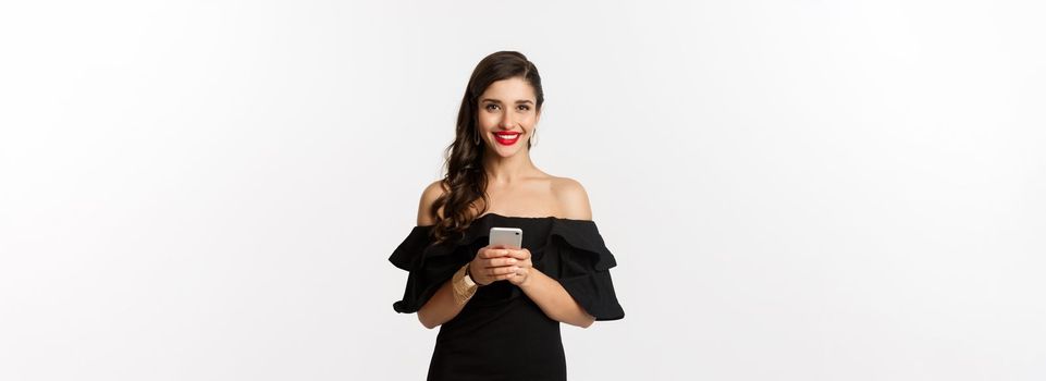 Online shopping concept. Attractive young woman in black dress, reading text message, using mobile phone and smiling, standing over white background.