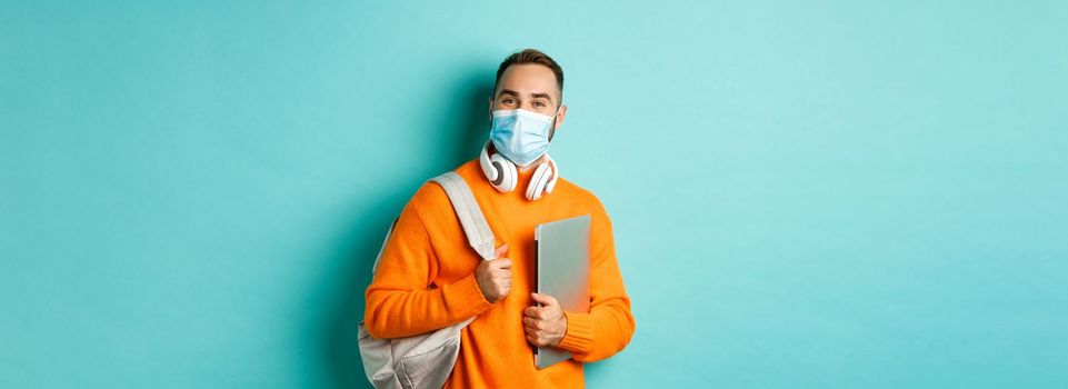 Handsome student in face mask, holding backpack and laptop, going work, standing over light blue background.