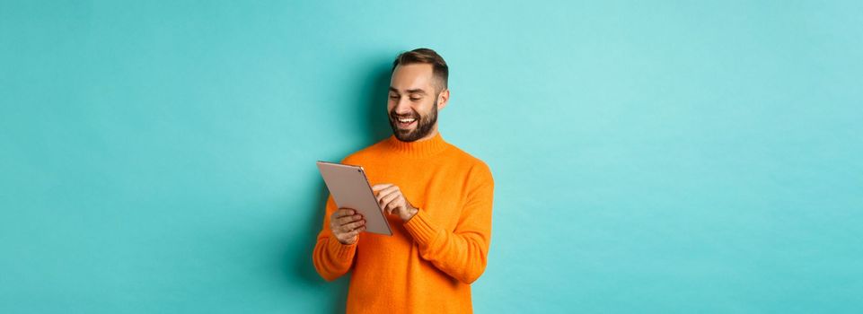Handsome adult man working on digital tablet and smiling, shopping online, standing over light blue background.
