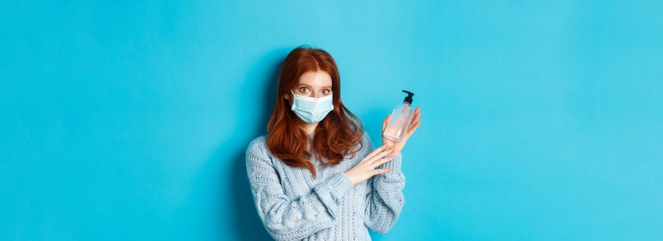 Winter, covid-19 and social distancing concept. Young redhead girl in face mask showing hand sanitizer, demonstrating antiseptic for disinfection, standing over blue background.