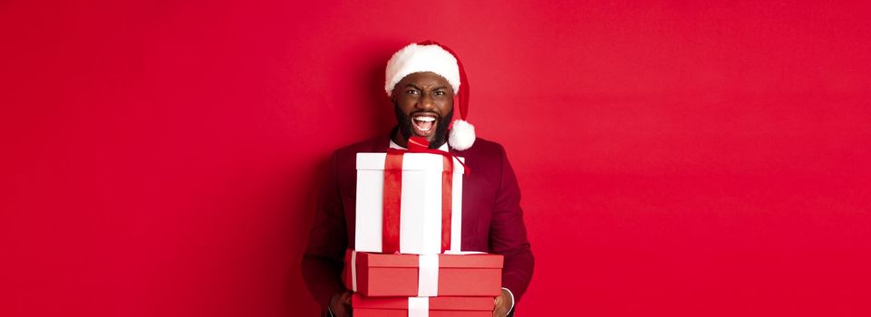 Christmas, New Year and shopping concept. Happy Black man in santa hat and blazer holding xmas presents, bring gifts and smiling, standing against red background.