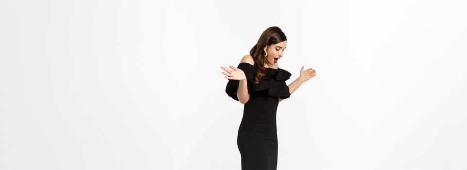 Full length view of woman in elegant dress and red lips, looking surprised, receive gifts on christmas holidays, standing with presents over white background.