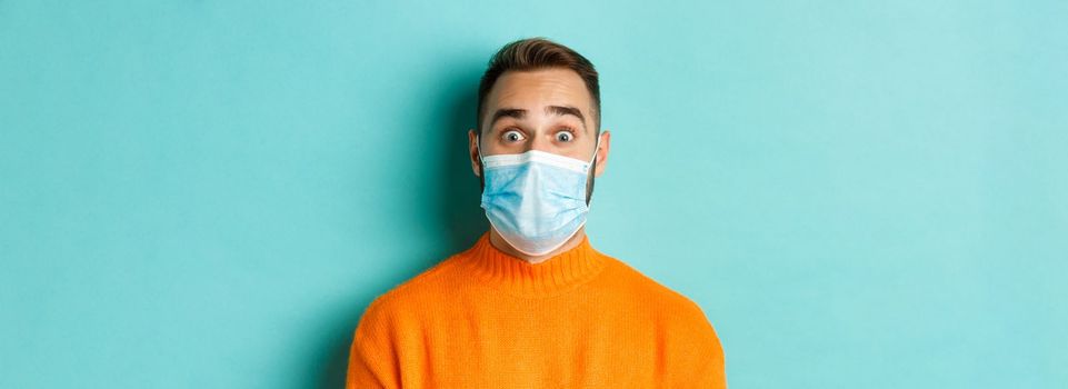 Covid-19, social distancing and quarantine concept. Close-up of young man in face mask staring at camera with surprised face, standing over turquoise background.