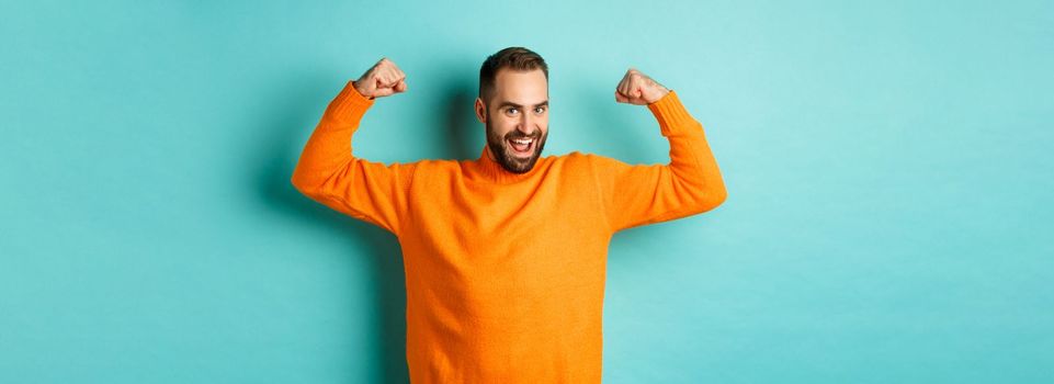 Young and strong man looking confident, flex biceps and show-off muscle strengths, smiling self-assured, standing over light blue background.