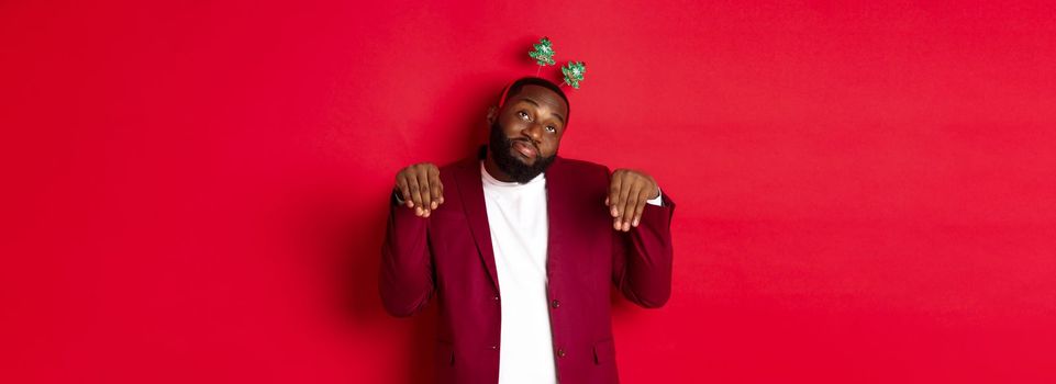 Merry Christmas. Silly and funny Black man in party headband, imitating bunny or cute puppy, standing over red background.