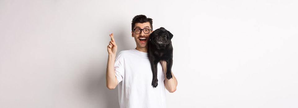 Hopeful smiling dog owner making a wish, holding cute black pug on shoulder and cross fingers for good luck, white background.