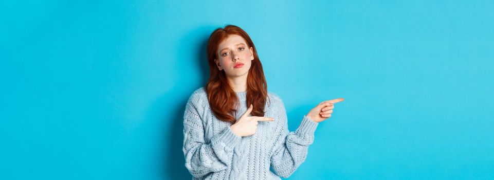 Moody teenage girl with red hair, pointing fingers left at logo, staring bothered and bored, standing over blue background.