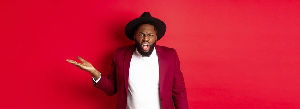 Angry and confused Black man arguing, shouting at camera and raising hand bothered, cant understand something, red background.