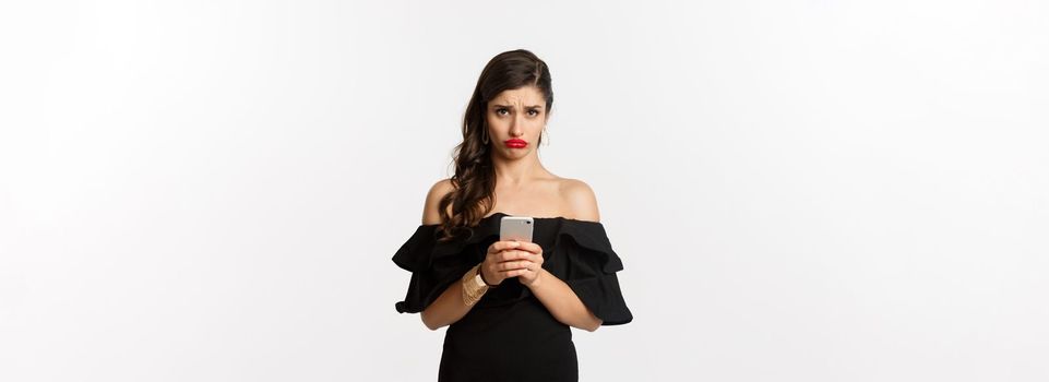 Sad and gloomy woman in black dress, sulking upset, using mobile phone and feeling disappointed, standing over white background.