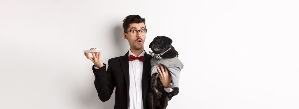 Handsome young hipster in suit and glasses holding cute black pug and pet food on plate, standing over white background.