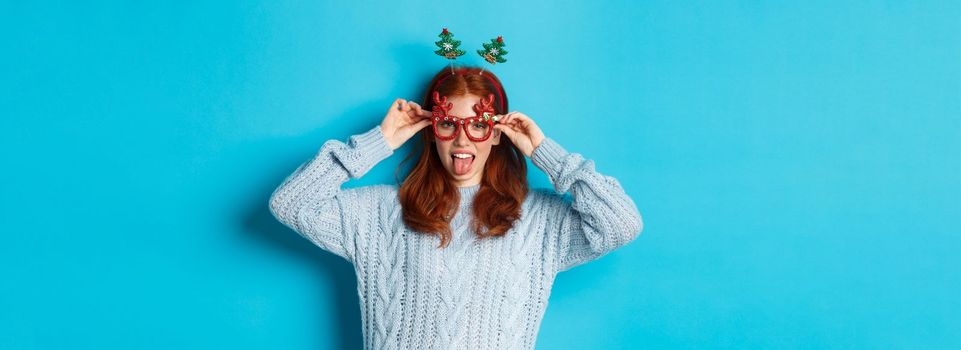 Winter holidays and Christmas sales concept. Beautiful redhead female model celebrating New Year, wearing funny party headband and glasses, smiling at camera.