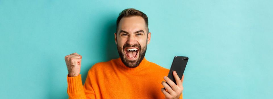 Image of man winning on mobile phone, making fist pump and rejoicing of victory, holding smartphone with satisfied face, turquoise background.