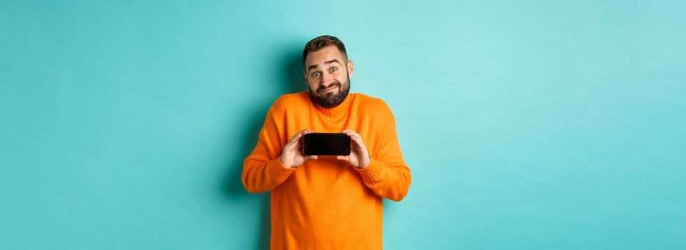 Clueless guy shrugging and showing mobile screen, indecisive emotion, standing in orange sweater over turquoise background.