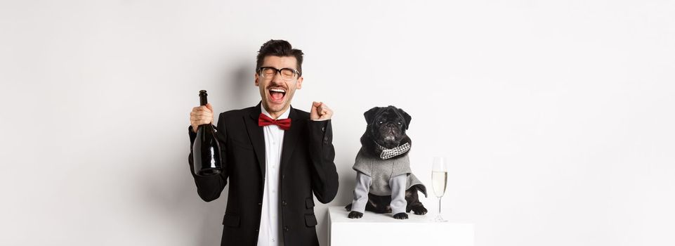 Pets, winter holidays and New Year concept. Happy young man celebrating Christmas with cute black dog wearing party costume, holding bottle champagne, white background.