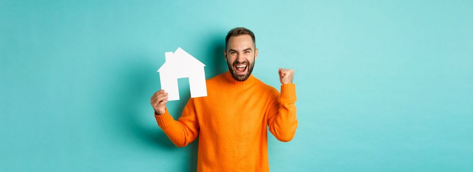 Real estate concept. Excited man saying yes, showing paper house maket and looking satisfied, standing in orange sweater over light blue background.