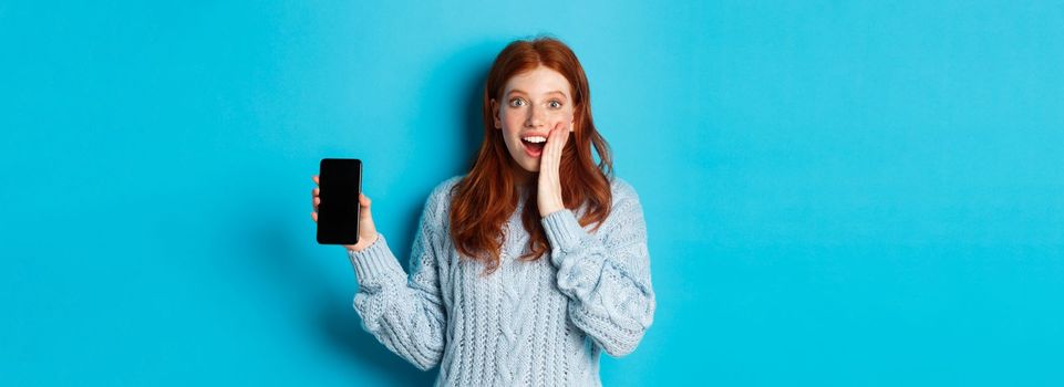Amazed redhead girl looking at camera, showing smartphone screen, demonstrating mobile application, standing over blue background.