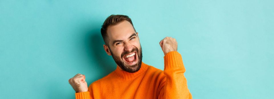 Image of handsome relieved man feeling satisfaction, rejoicing of winning or achievement, making fist pump and saying yes, standing over turquoise background.