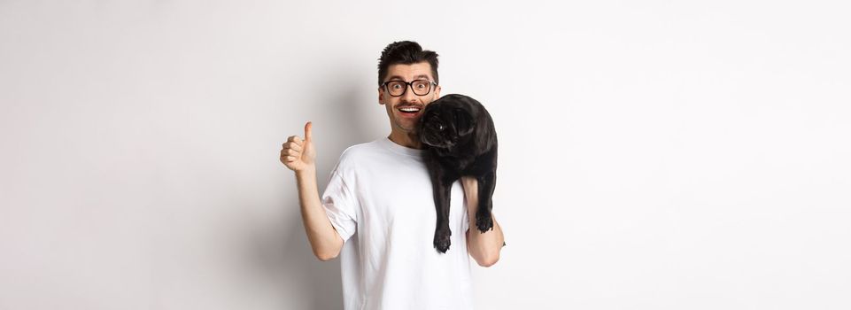 Happy and satisfied dog owner showing thumb-up, holding cute black pug on shoulder, recommending pet products, standing over white background.