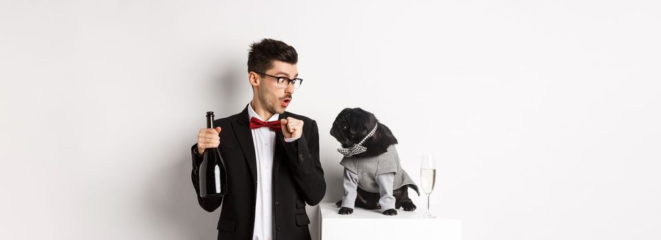 Pets, winter holidays and New Year concept. Happy young man celebrating Christmas with cute black dog wearing party costume, puppy looking at owner, white background.