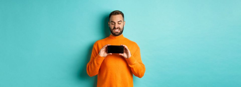 Image of man cringe at something on mobile screen, staring disgusted at display, showing smartphone, standing over light blue background.