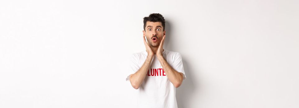Shocked man in volunteer t-shirt gasping, staring at camera in awe, standing over white background.