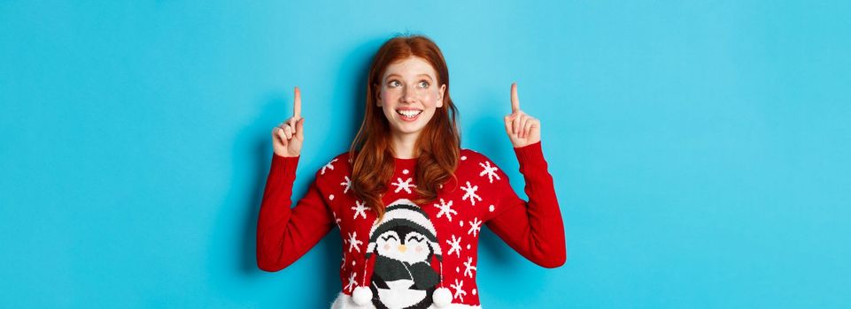 Winter holidays and celebration concept. Cheerful teen girl with red hair, looking dreamy at logo, pointing fingers up, showing advertisement, standing over blue background.