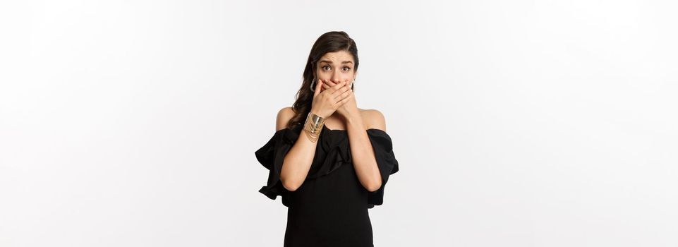 Fashion and beauty. Attractive woman in black dress cover mouth and gasping shocked, staring at camera worried, standing over white background.