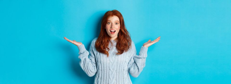 Image of redhead young woman spread hands sidewas, looking with disbelief and amazement, hear news, standing over blue background.