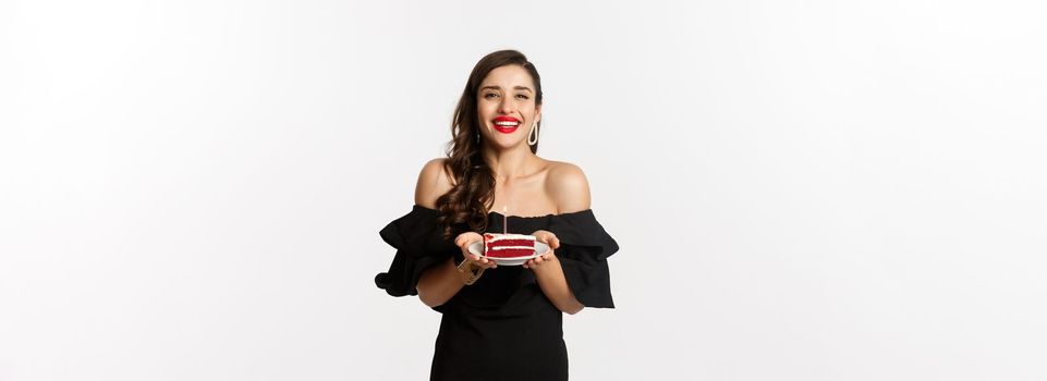 Celebration and party concept. Happy gorgeous woman having birthday, holding b-day cake and smiling, making wish, standing in black dress with makeup.