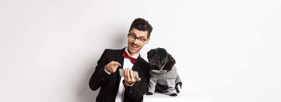 Happy dog owner showing something to pet on mobile phone, man and pug wearing fancy costumes, standing over white background.