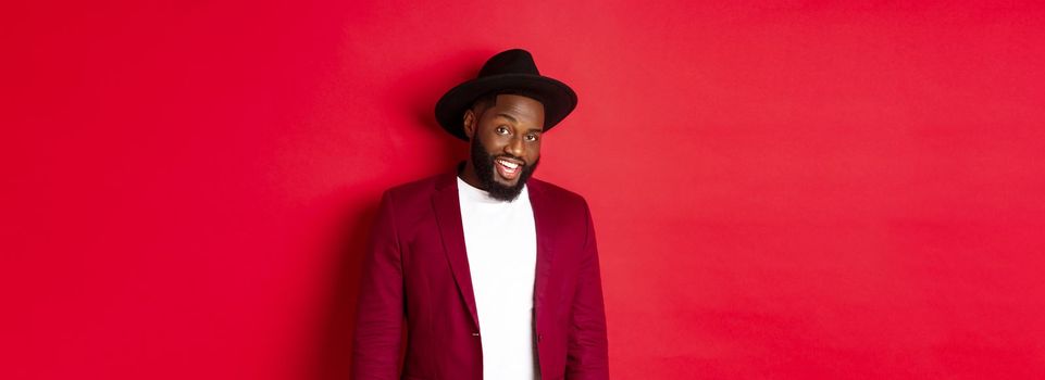 Christmas shopping and people concept. Handsome bearded african american guy looking at camera, smiling confident, wearing party clothes, red background.