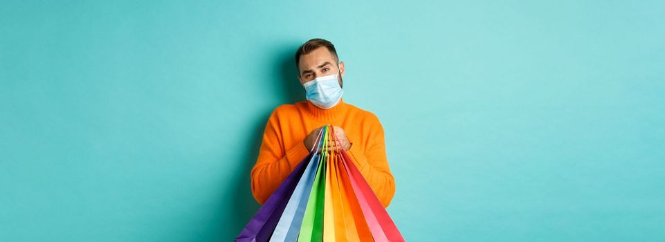 Covid-19, pandemic and lifestyle concept. Displeased and reluctant man in medical mask, holding shopping bags and complaining, standing over turquoise background.