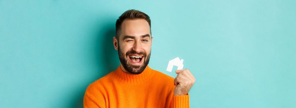 Real estate concept. Happy young man searching for home rent, holding house paper maket and smiling, standing over blue background.