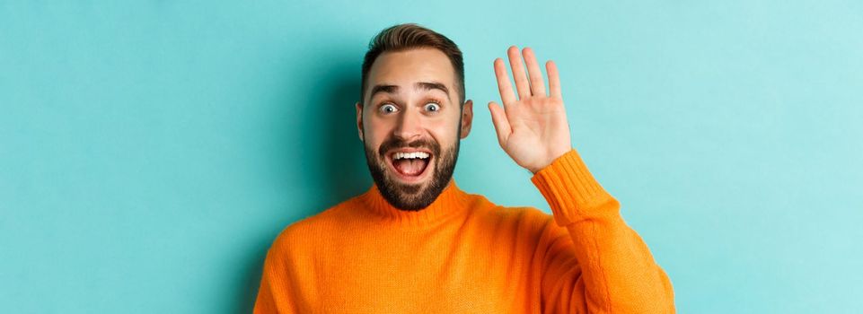 Handsome man waving hand to say hello, giving high five, standing over light blue background.