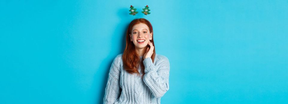 Winter holidays and Christmas sales concept. Beautiful redhead female model celebrating New Year, wearing funny party headband and sweater, smiling at camera.