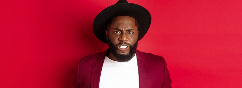 Tensed african american man clench teeth and frowning, looking at something uncomfortable and cringe, standing over red background.