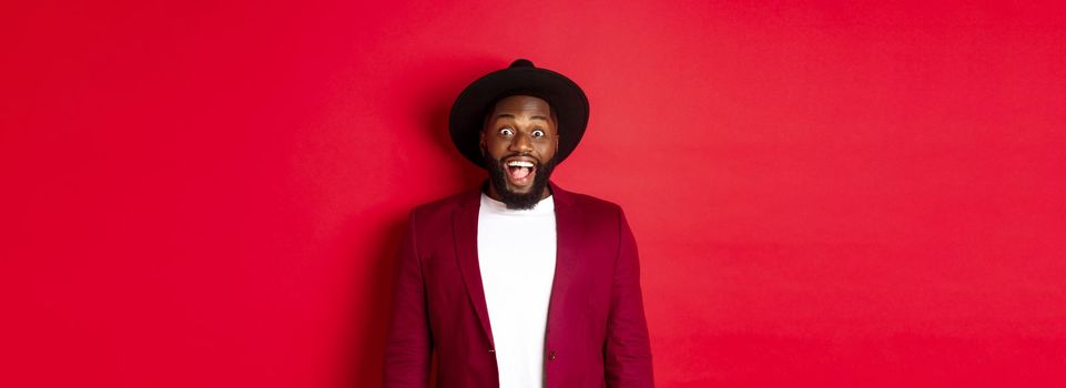 Fashion and party concept. Cheerful african american man in jacket and fancy hat, looking amazed and happy at camera, checking out new year promo, red background.