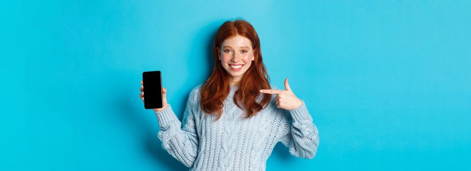 Satisfied redhead girl pointing at phone screen, showing smartphone app or online promo and smiling, standing in sweater against blue background.