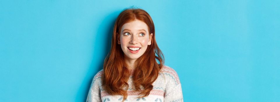 Close-up of dreamy redhead girl in winter sweater looking left, smiling and staring at promo offer, standing against blue background.