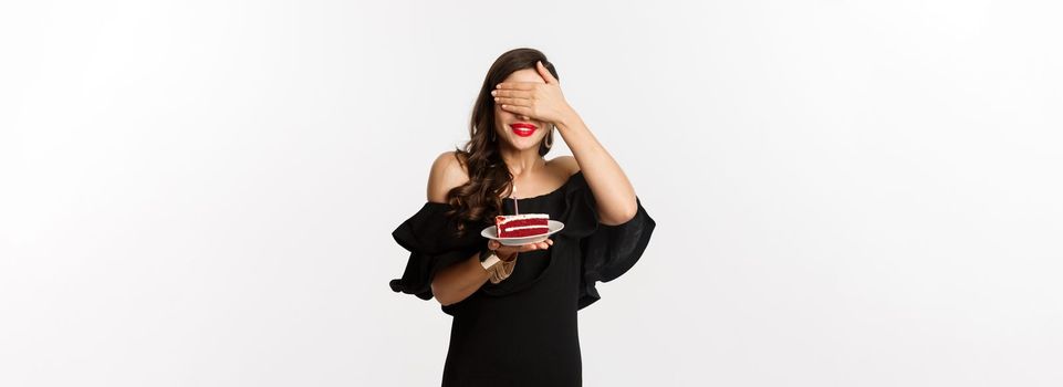 Celebration and party concept. Happy birthday girl in black dress, red lipstick, close eyes and making wish on b-day cake, standing over white background.