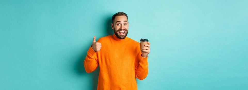 Handsome satisfied man showing thumbs-up, like coffee, smiling pleased, standing over turquoise background. Copy space