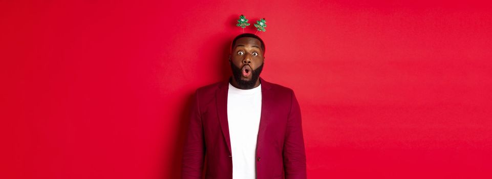Merry Christmas. Happy african american man celebarting New Year, wearing party headband and making funny faces , standing over red background.