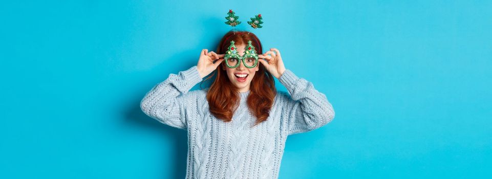 Winter holidays and Christmas sales concept. Beautiful redhead female model celebrating New Year, wearing funny party headband and glasses, smiling silly, blue background.