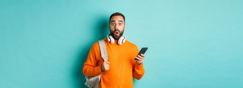 Caucasian man with headphones and backpack staring at camera surprised after reading phone message, standing over turquoise background.