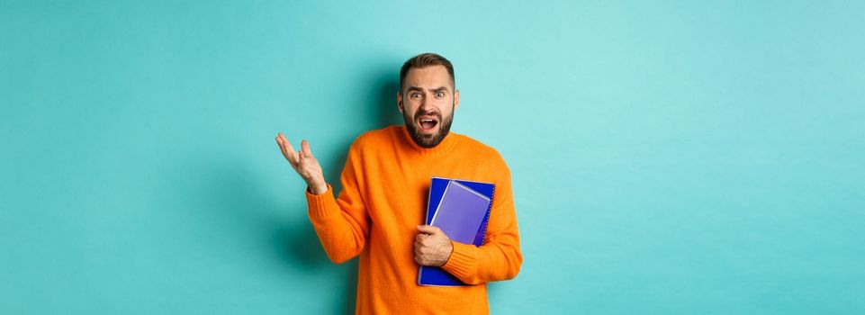 Education. Confused and disappointed man arguing, holding notebooks and complaining, standing over light blue background.