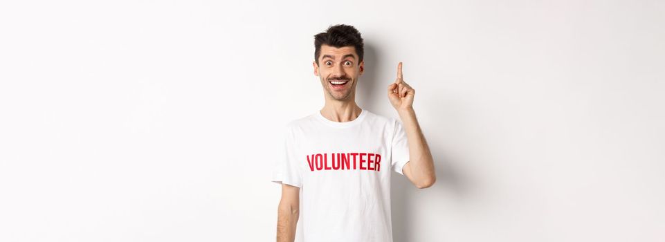 Handsome young man in volunteer t-shirt having an idea, raising finger and saying suggestion, pointing up, standing over white background.