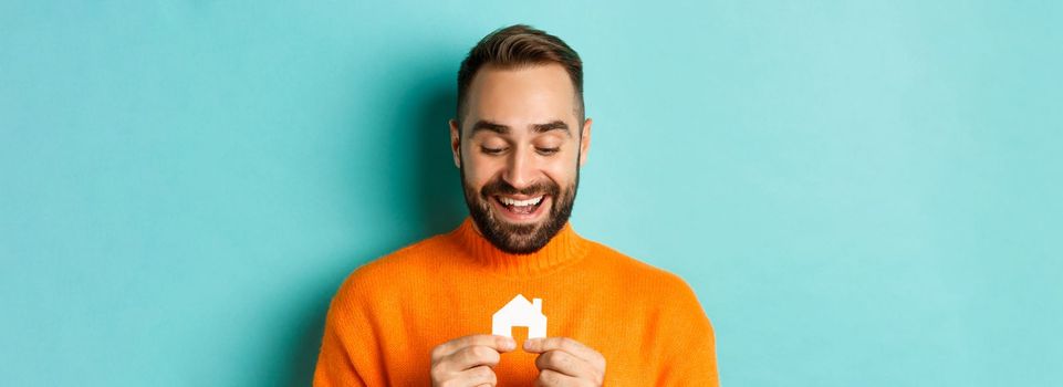 Real estate concept. Happy young man searching for home rent, holding house paper maket and smiling, standing over blue background.