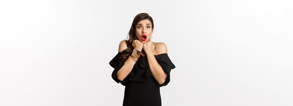 Portrait of glamour woman looking scared and shocked at camera, staring at something with fear, standing in black dress against white background.